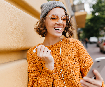 A woman wearing an
            orange sweater, orange sunglasses, and a gray hat, holding a phone
            in one hand and earphones in the other.